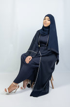 A woman seated wearing a full-length kimono in shades of blue with detailed stitching, paired with a matching blue headscarf.